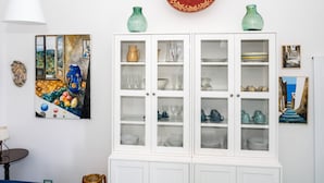 Ein Zimmer mit einem weißen Schrank, der Glaswaren und Geschirr präsentiert, einem Gemälde einer Obstschale und einer Pendelleuchte.