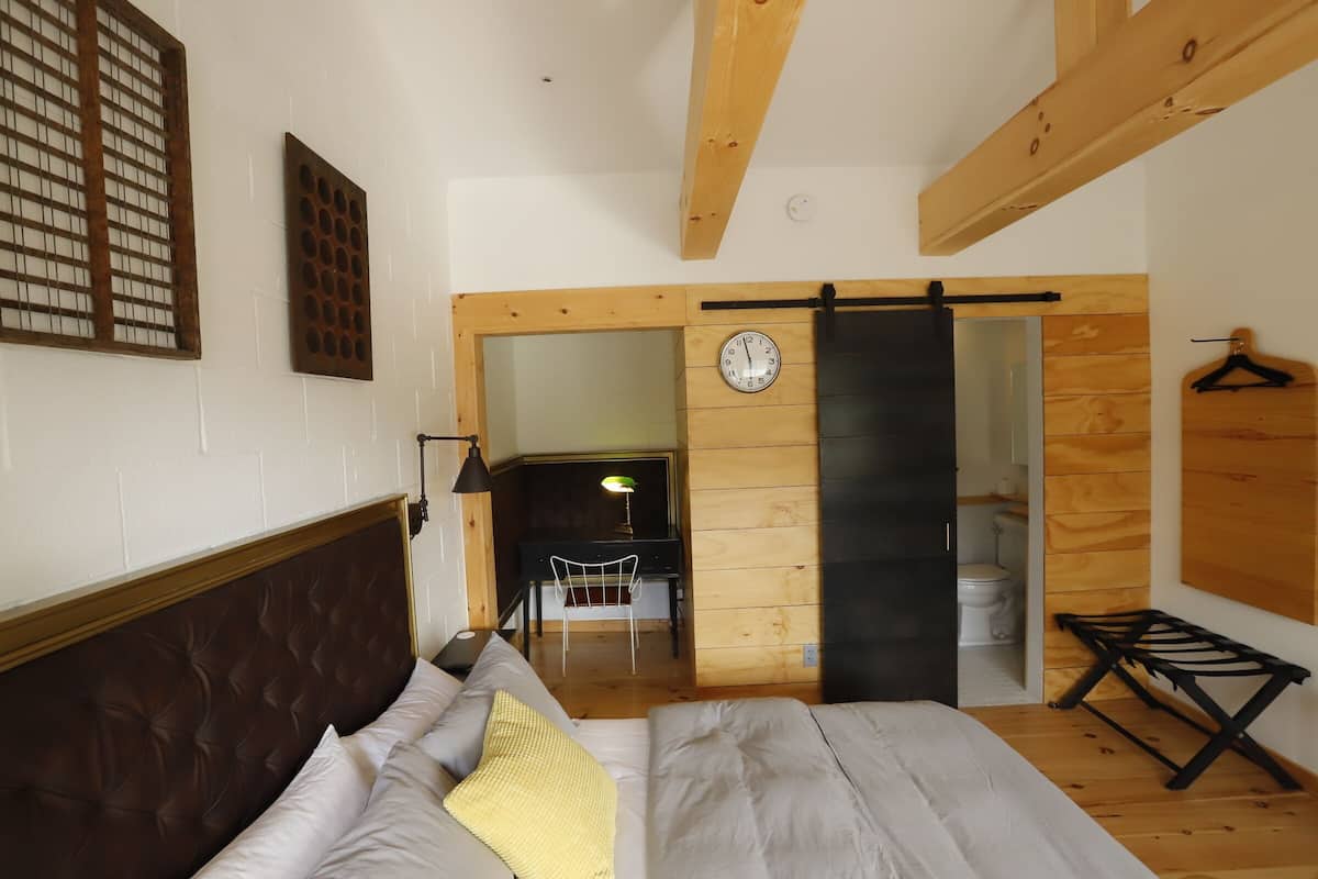 Quarto com cama, cabeceira de madeira, relógio na parede e porta de madeira que dá para outro cômodo.