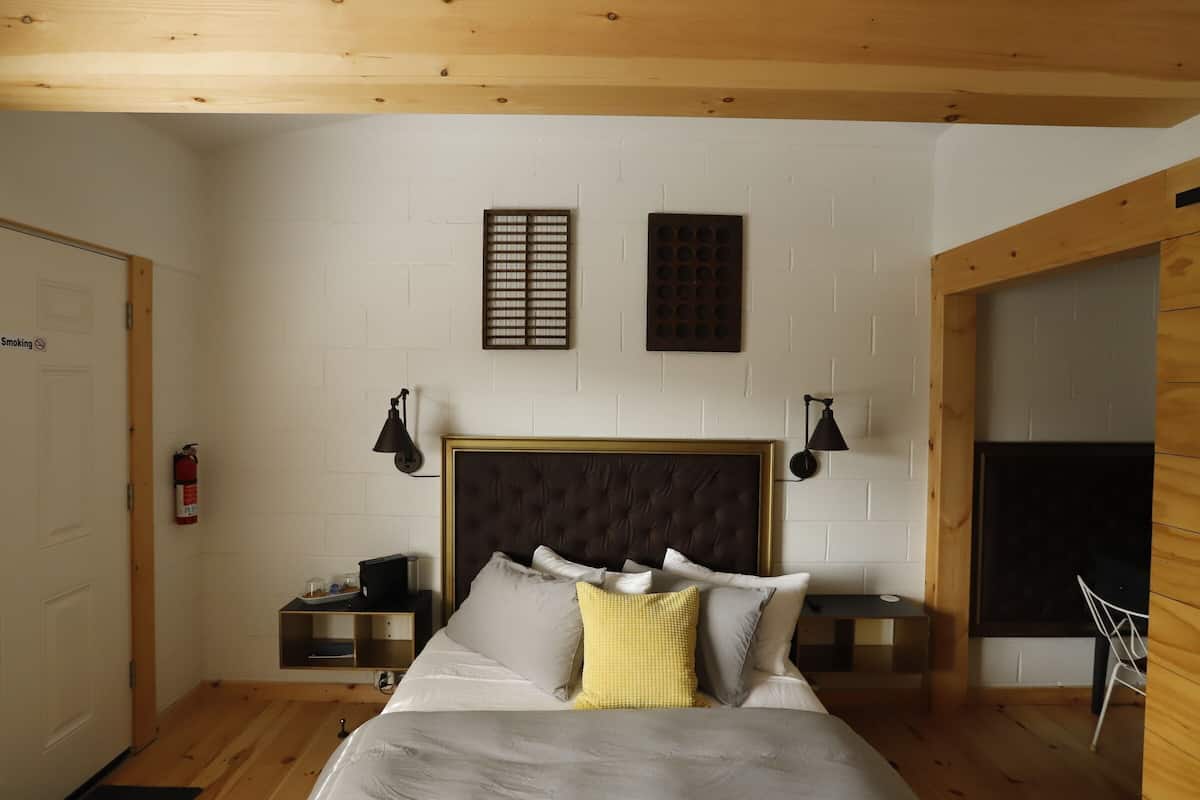 Quarto com cabeceira estofada, cama com várias almofadas, mesas de cabeceira, extintor de incêndio e uma janela.