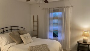Quarto com cama, almofadas, luminária de cabeceira, janela com cortinas leves e uma escada de madeira.