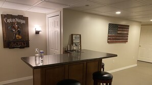 Uma área de bar com balcão de madeira, dois bancos, um cartaz de Captain Morgan e uma bandeira americana na parede.