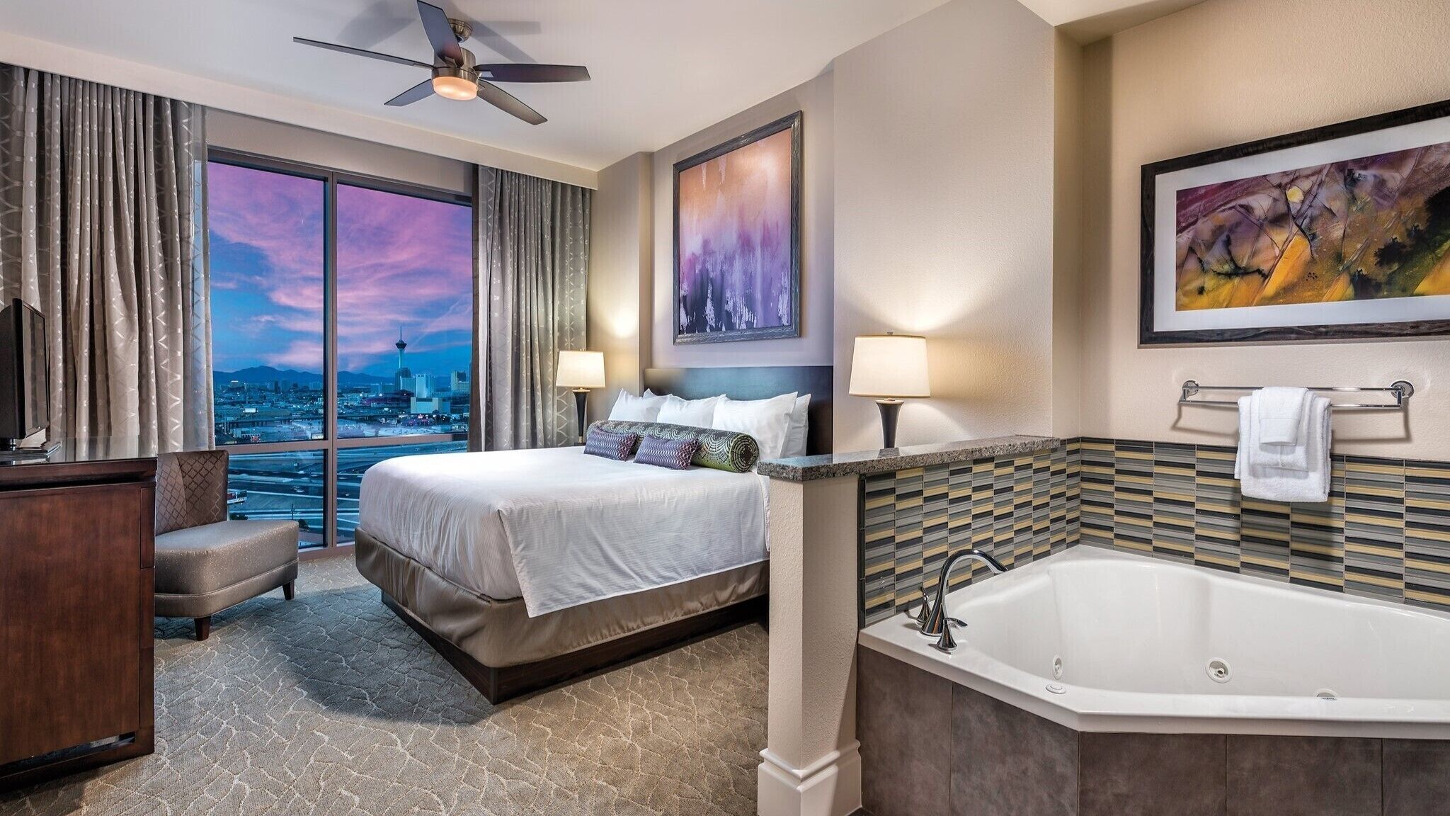 A hotel room with a large bed, a sitting area, a bathtub, and a city view through large windows.