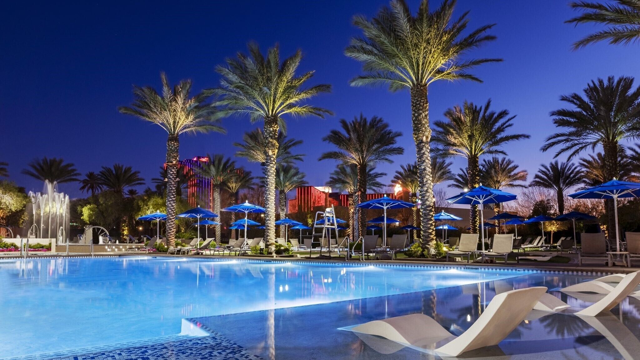A pool area with lounge chairs and umbrellas, palm trees, and a fountain.