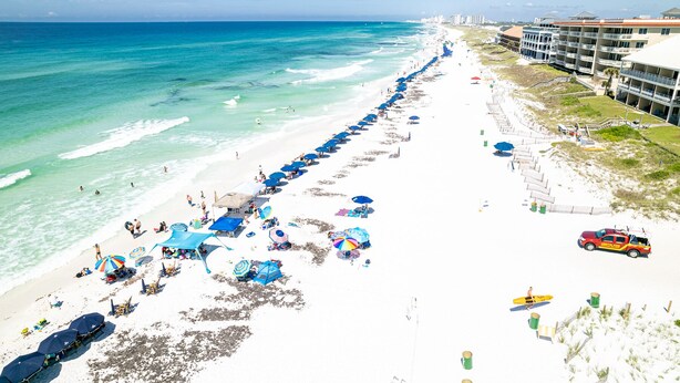 Una playa con una fila de sombrillas y sillas azules, una orilla arenosa y un edificio con balcones con vistas a la playa.