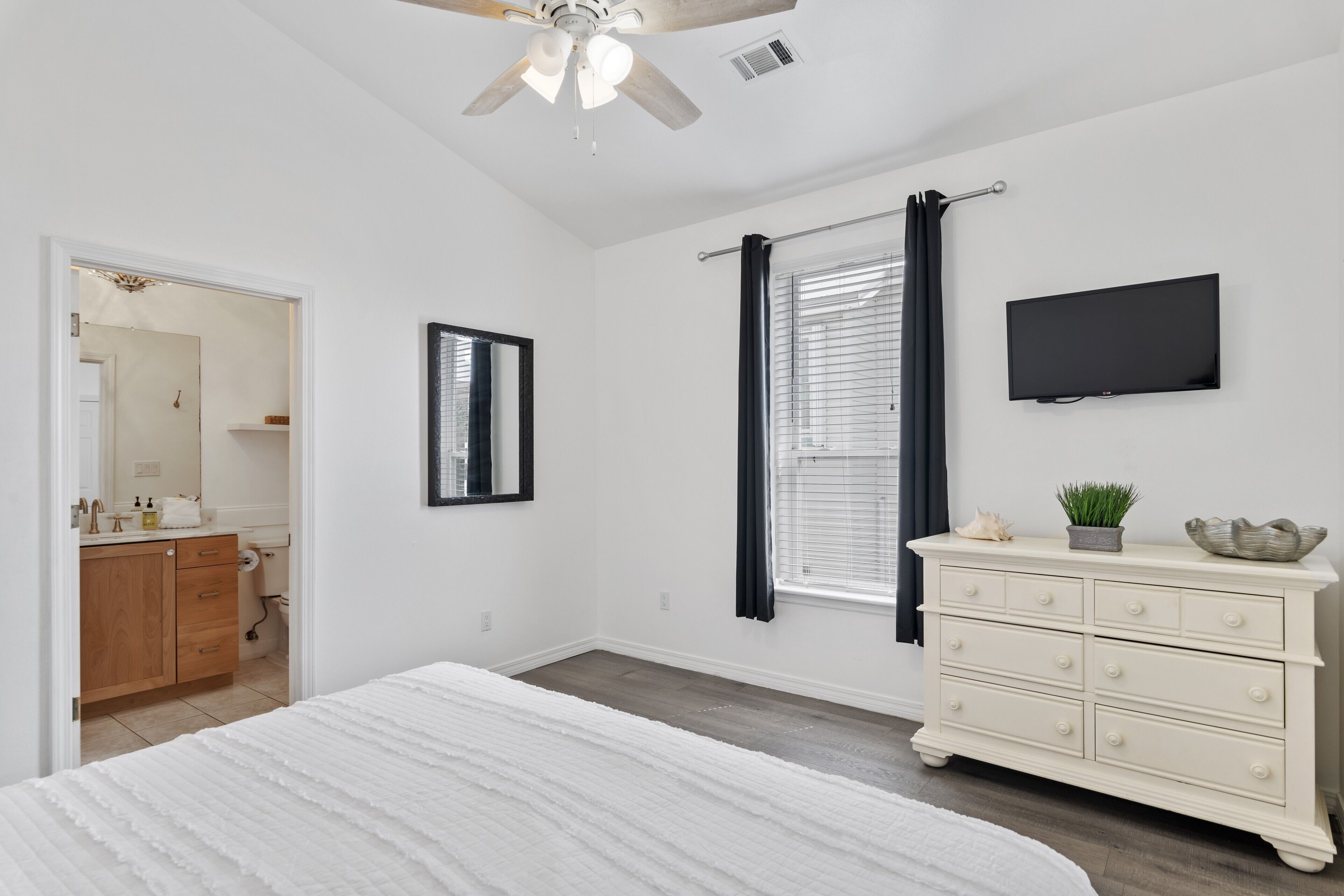 Une chambre à coucher avec un lit, une commode, un téléviseur et une fenêtre avec des rideaux.