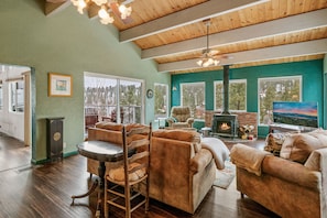 A spacious living room with a fireplace, wooden ceiling, and large windows offering a view of the outdoors.