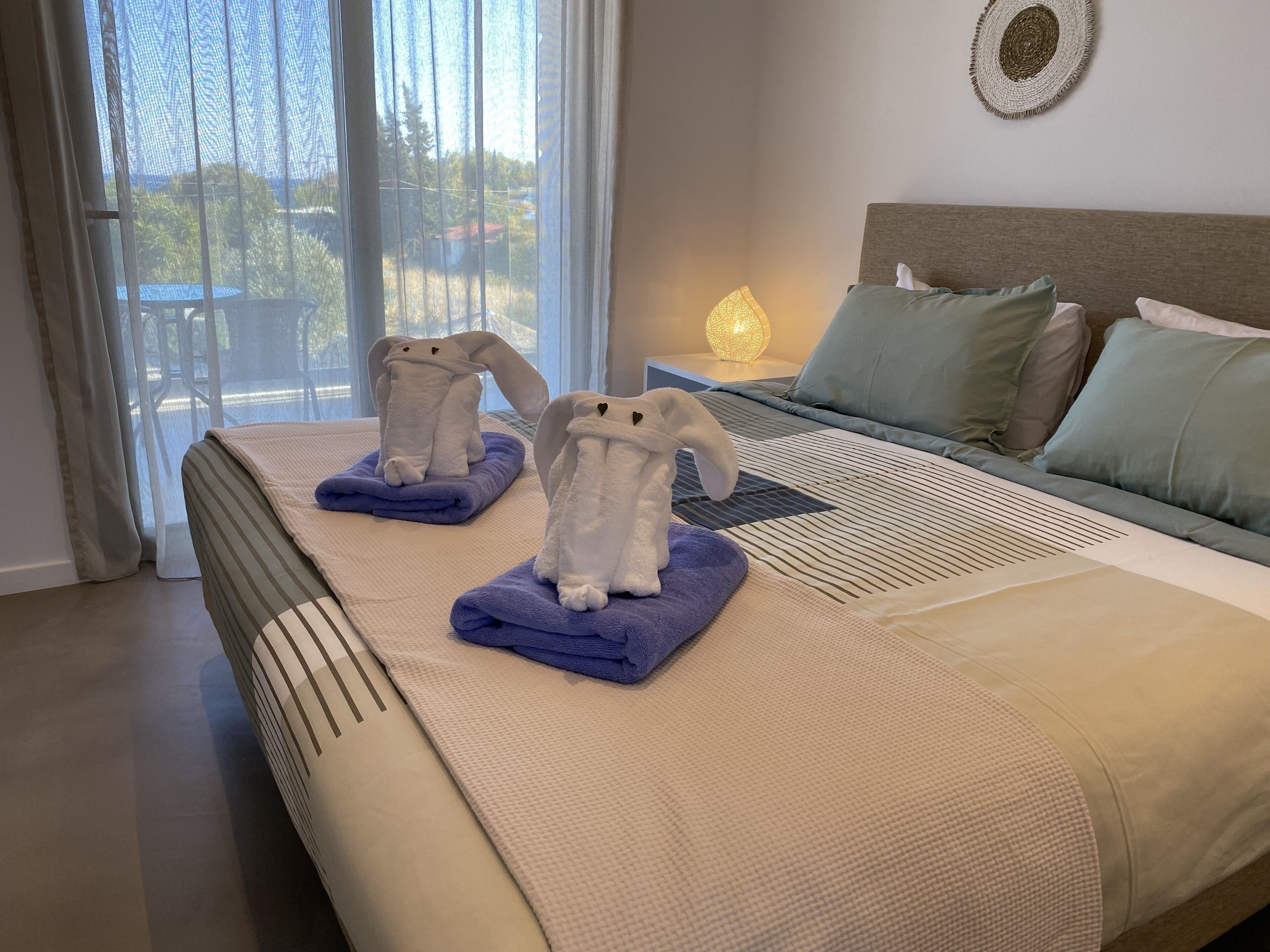 A hotel room with a bed, two towels shaped like animals, a nightstand with a lamp, and a view through the window.