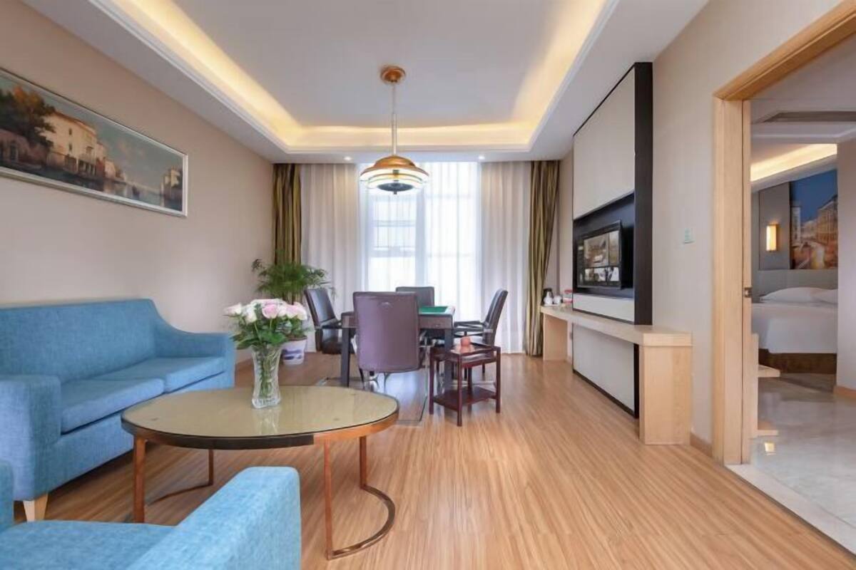 Ein modernes Hotelzimmer mit einem blauen Sofa, einem kleinen runden Tisch mit Blumenvase, einem Schreibtisch mit Computer und einem an der Wand montierten Fernseher.