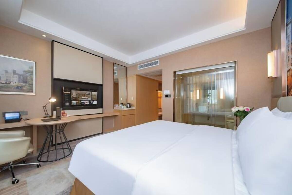 Ein Hotelzimmer mit einem großen Bett, einem Schreibtisch mit Computer, einem Stuhl, einem Fernseher und einer Glastür zum Badezimmer.