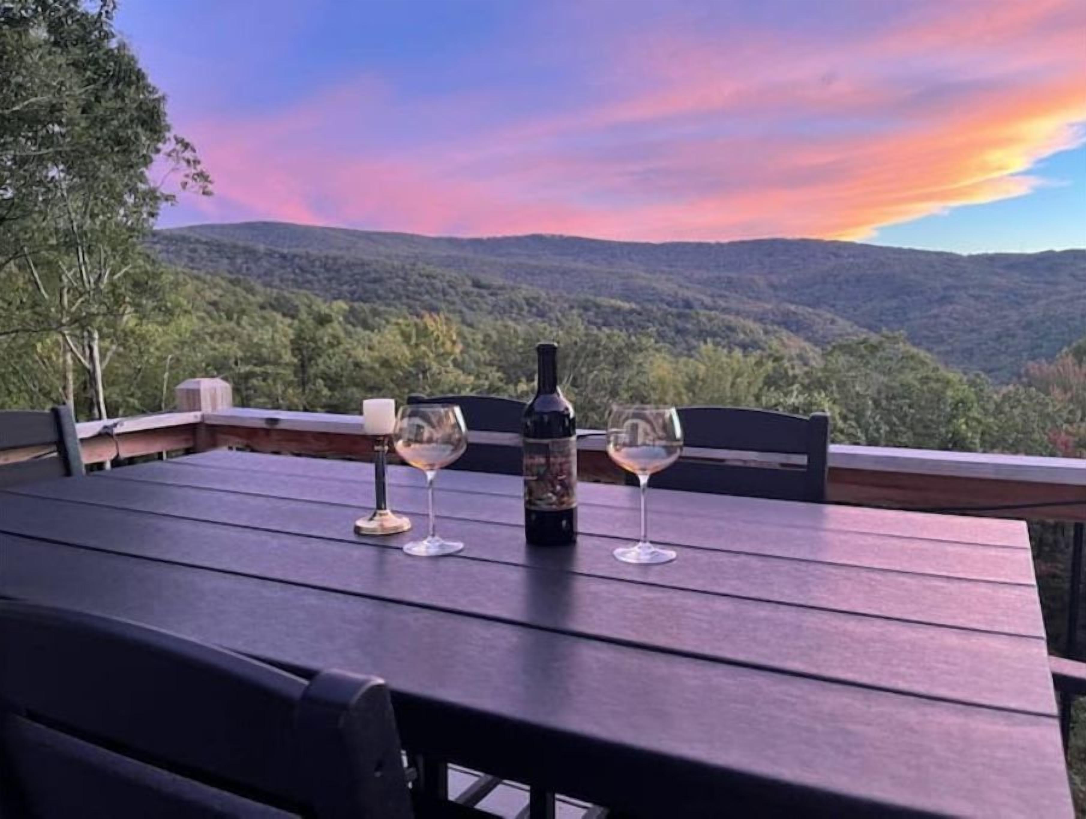 A table with two wine glasses and a bottle, set against a scenic view of a forested mountain range.
