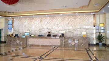 A hotel reception area with a marble counter, a receptionist, and a large plant.