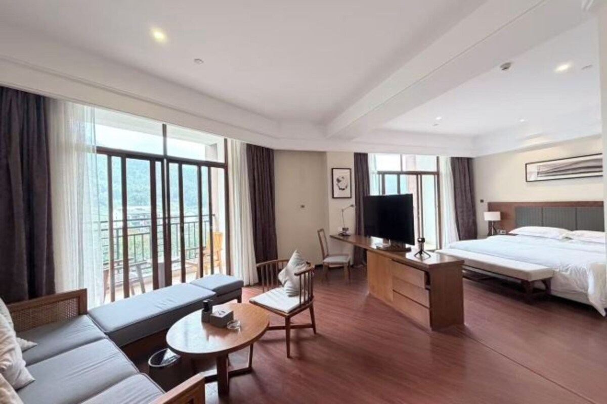 A hotel room with a bed, a sofa, a desk, a TV, and large windows.