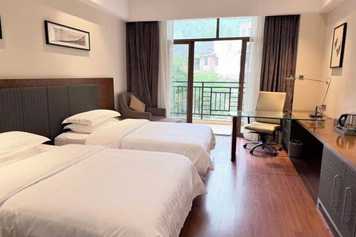 A hotel room with two beds, a desk with a chair, a window with curtains, and a balcony view.