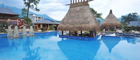 A swimming pool with thatched-roof structures, clear blue water, and a mountainous backdrop.