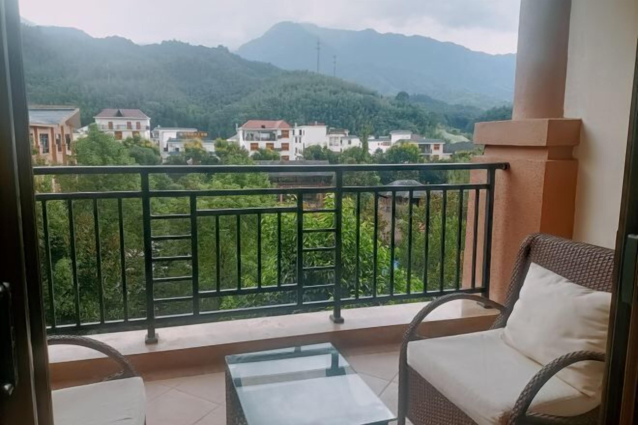 A balcony with a glass table and wicker chair, overlooking a mountainous area with buildings.