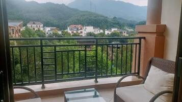 A balcony with a glass table and wicker chair, overlooking a mountainous area with buildings.