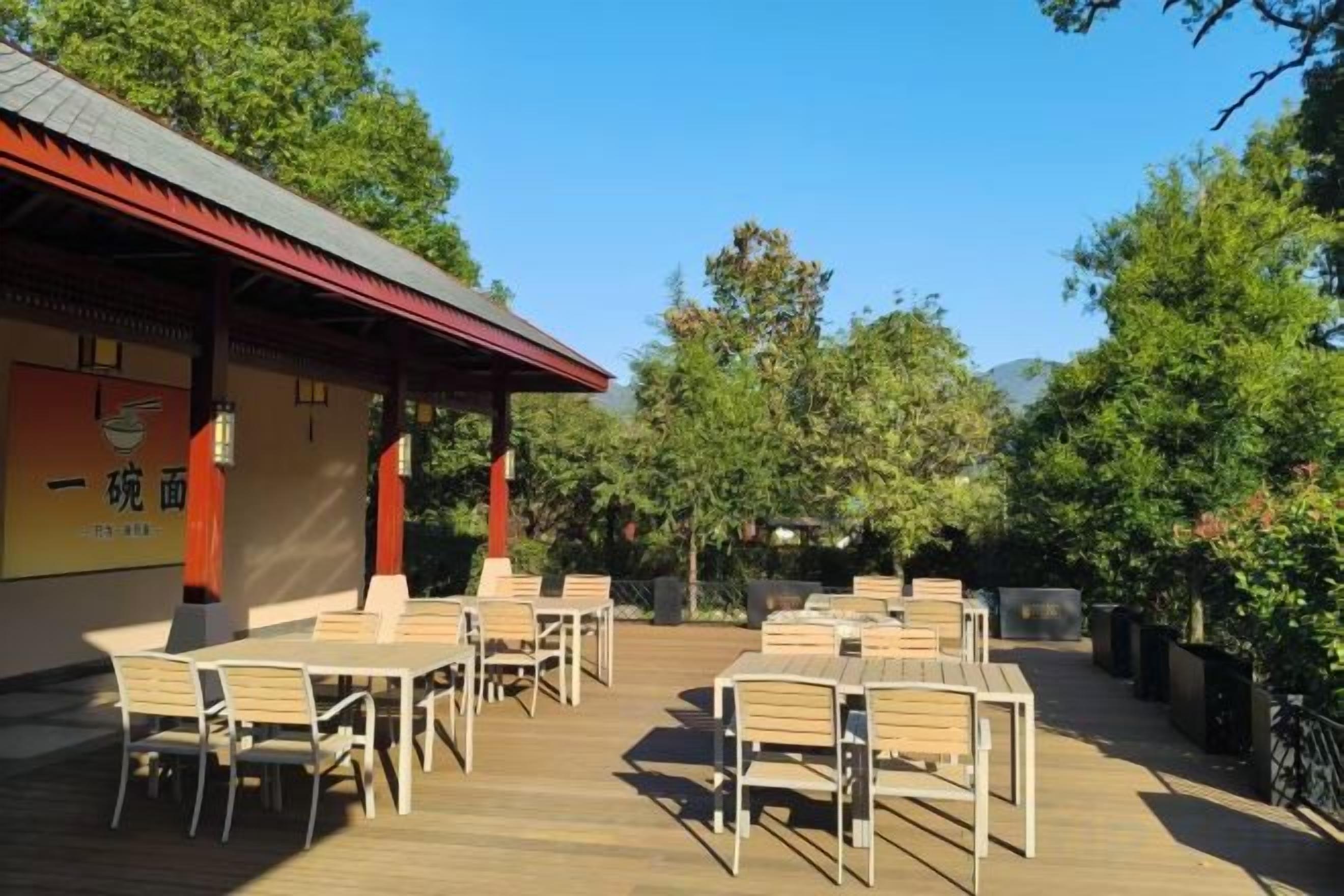 An outdoor seating area with tables and chairs, a building with signage, and trees in the background.