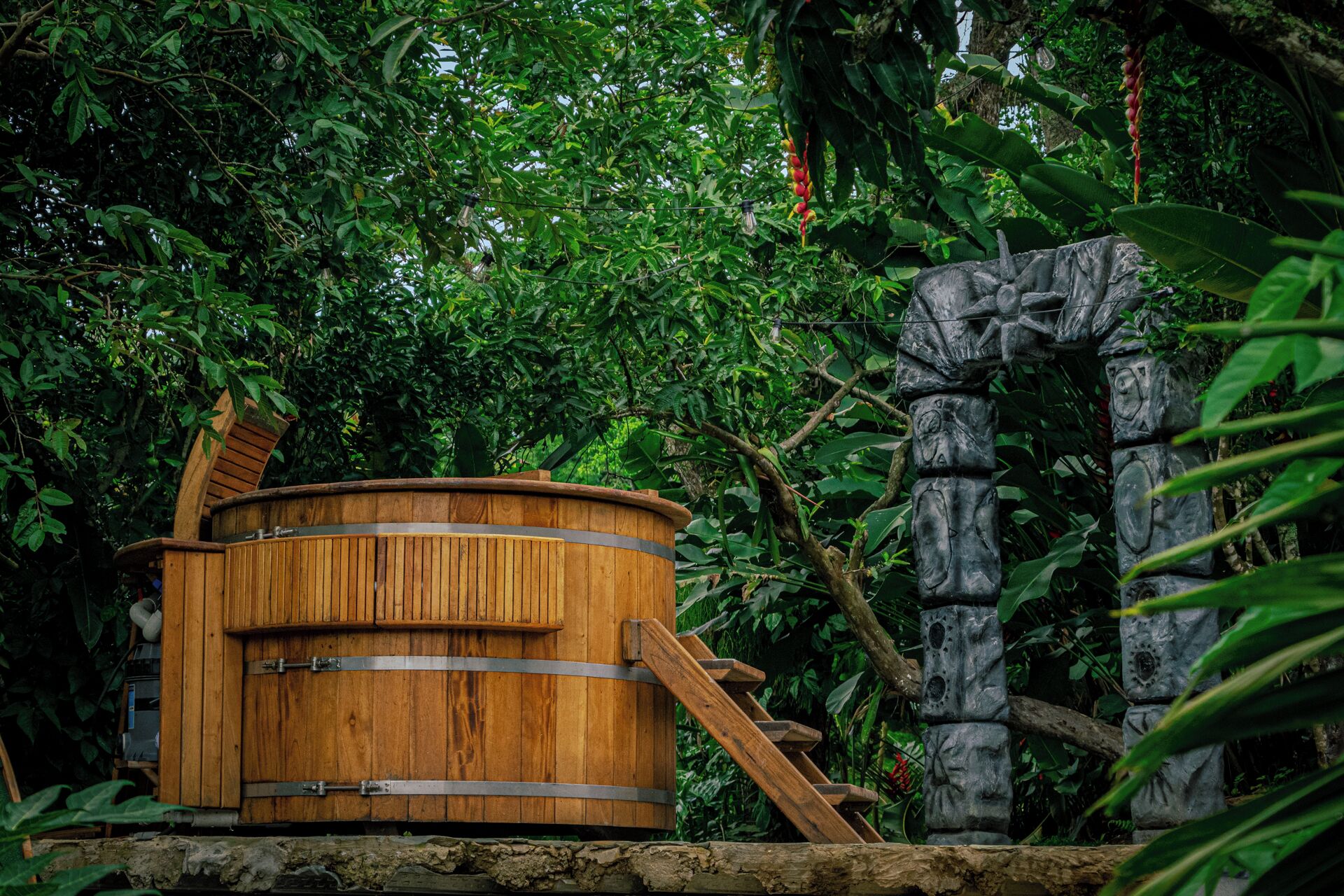 A wooden hot tub with metal bands, set against a backdrop of lush greenery and a stone structure resembling a totem.