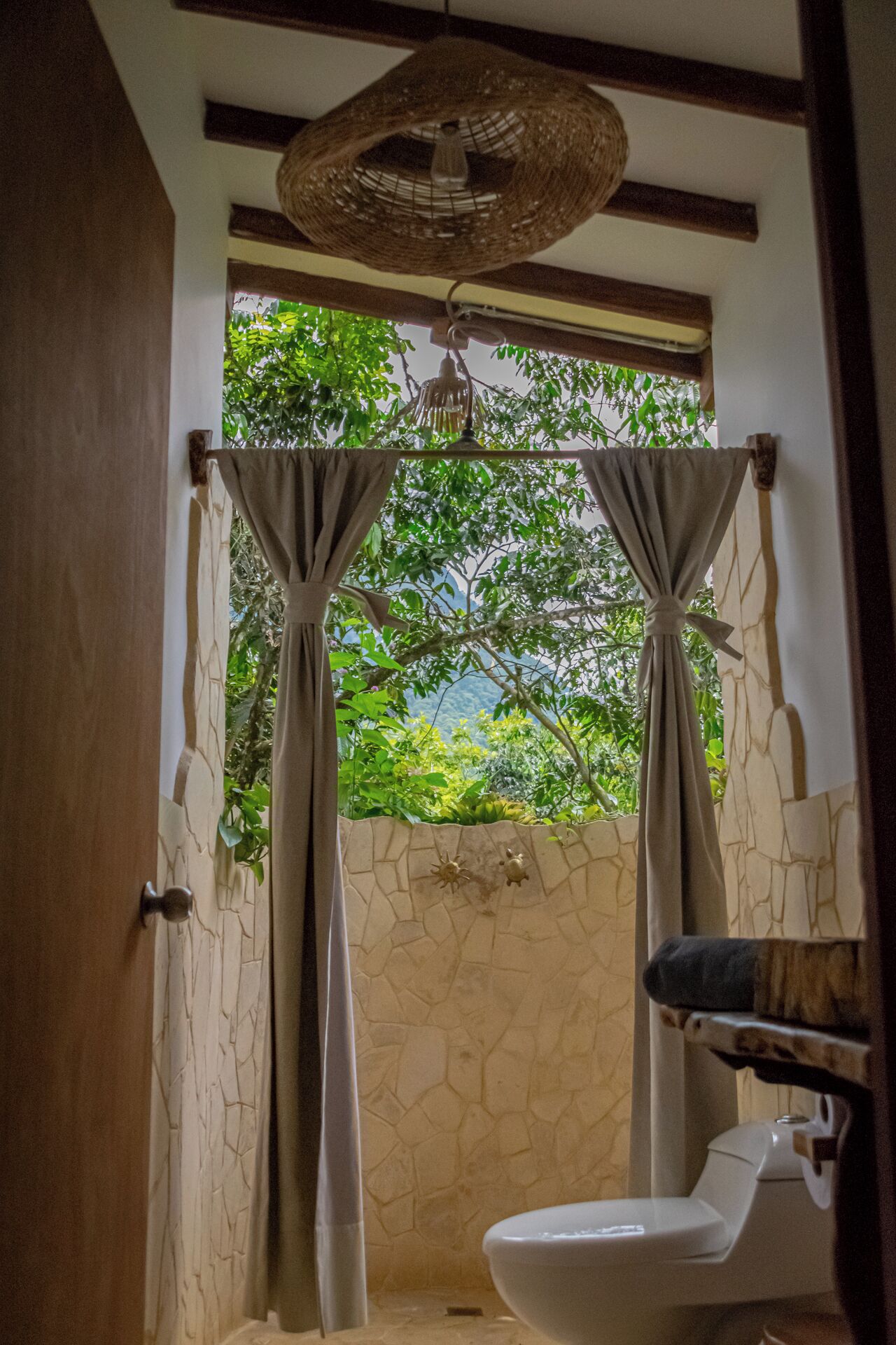 A bathroom with a stone wall, a white toilet, and a shower area with a woven ceiling light fixture.
