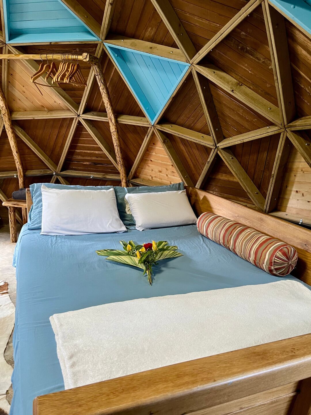 A wooden ceiling with a geometric pattern, a bed with blue bedding, white pillows, a red rose, and a striped pillow.