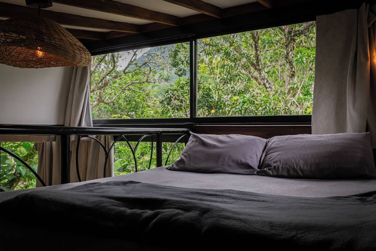 A bed with a dark blanket and pillows, a woven light fixture, and a window with curtains overlooking a forested area.