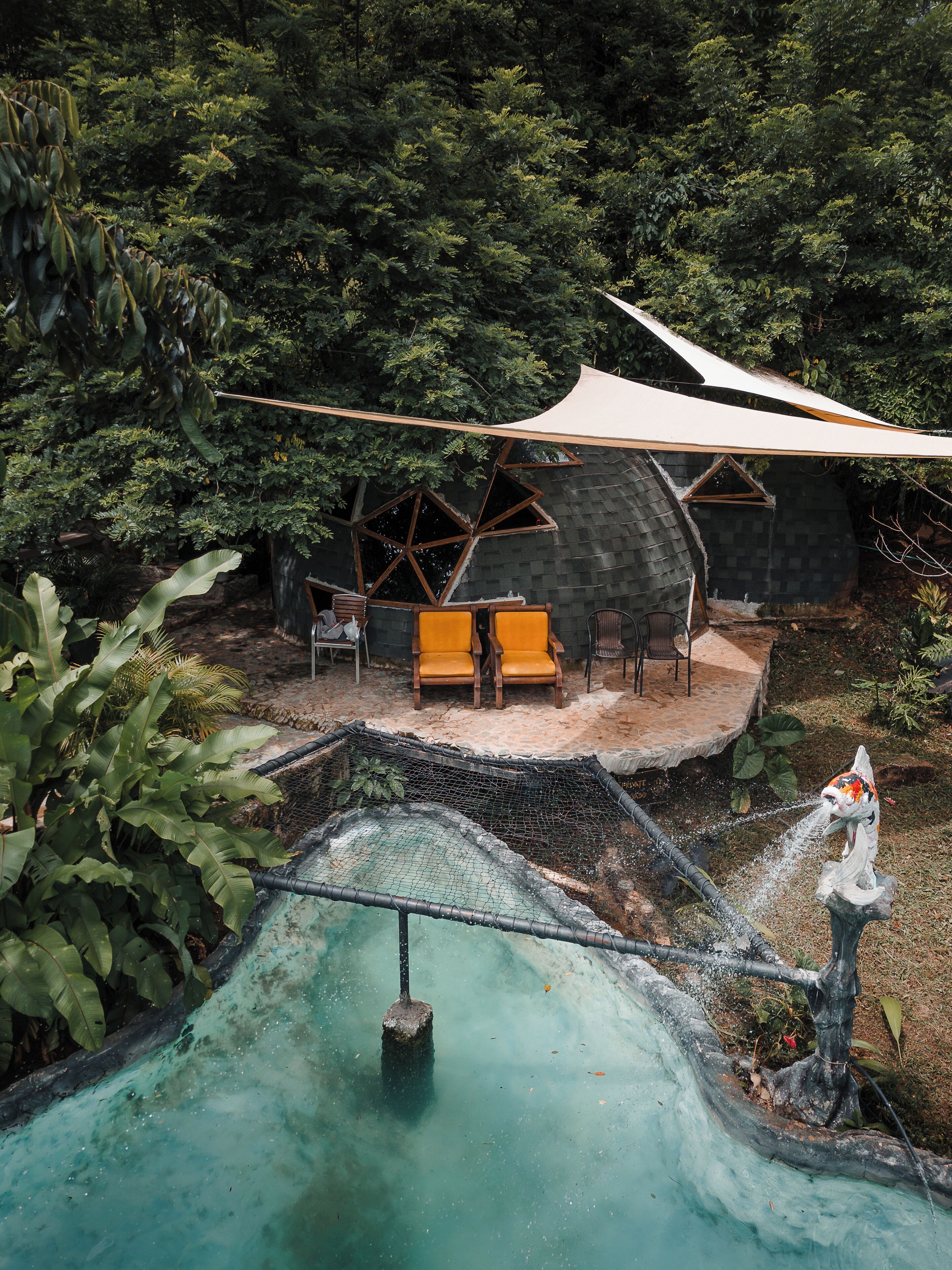 Una casa con techo inclinado, rodeada de un bosque denso. Cuenta con una piscina con cascada y un pequeño puente.