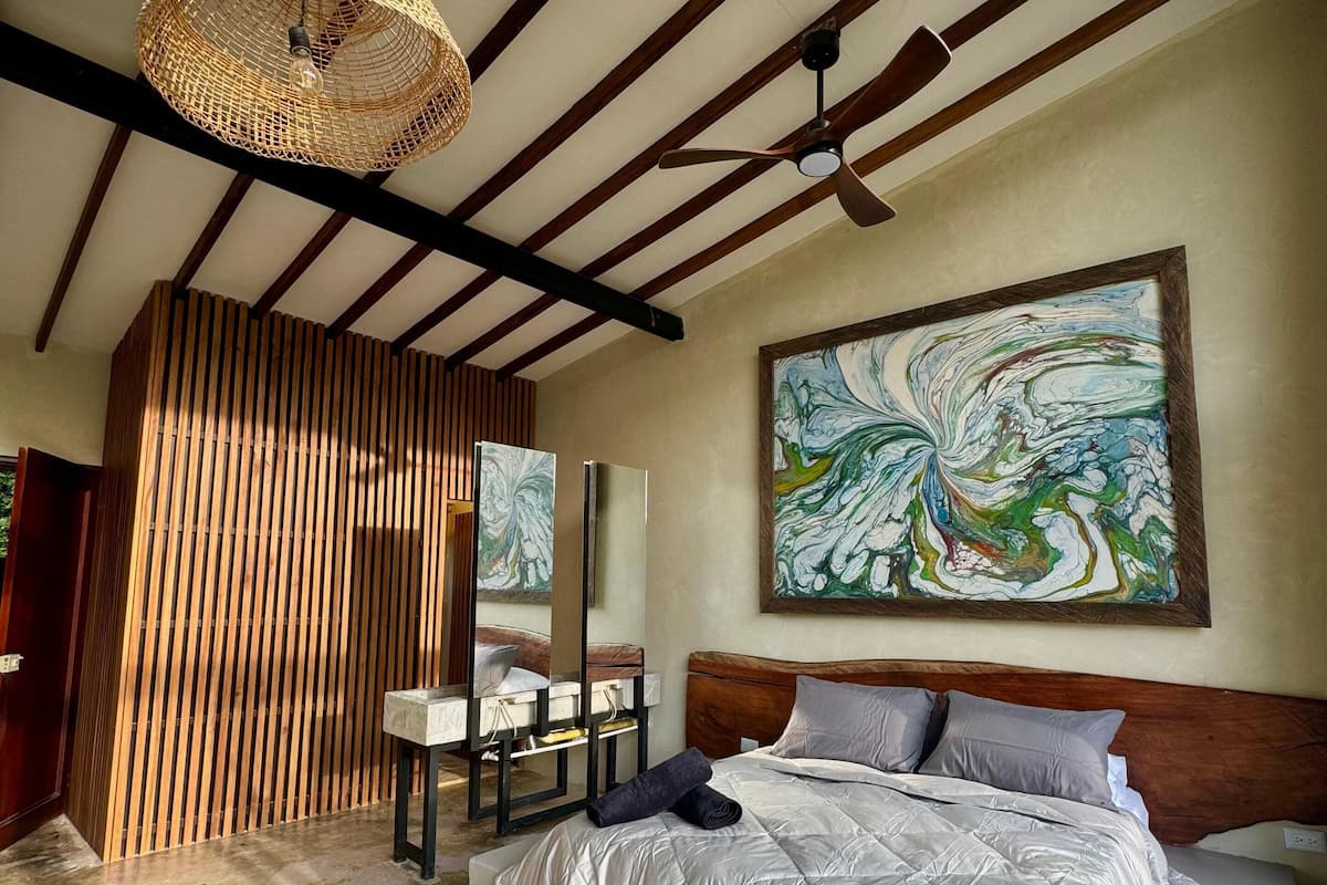 A modern bedroom with a bed, a ceiling fan, a large abstract painting, and a wooden slat partition.