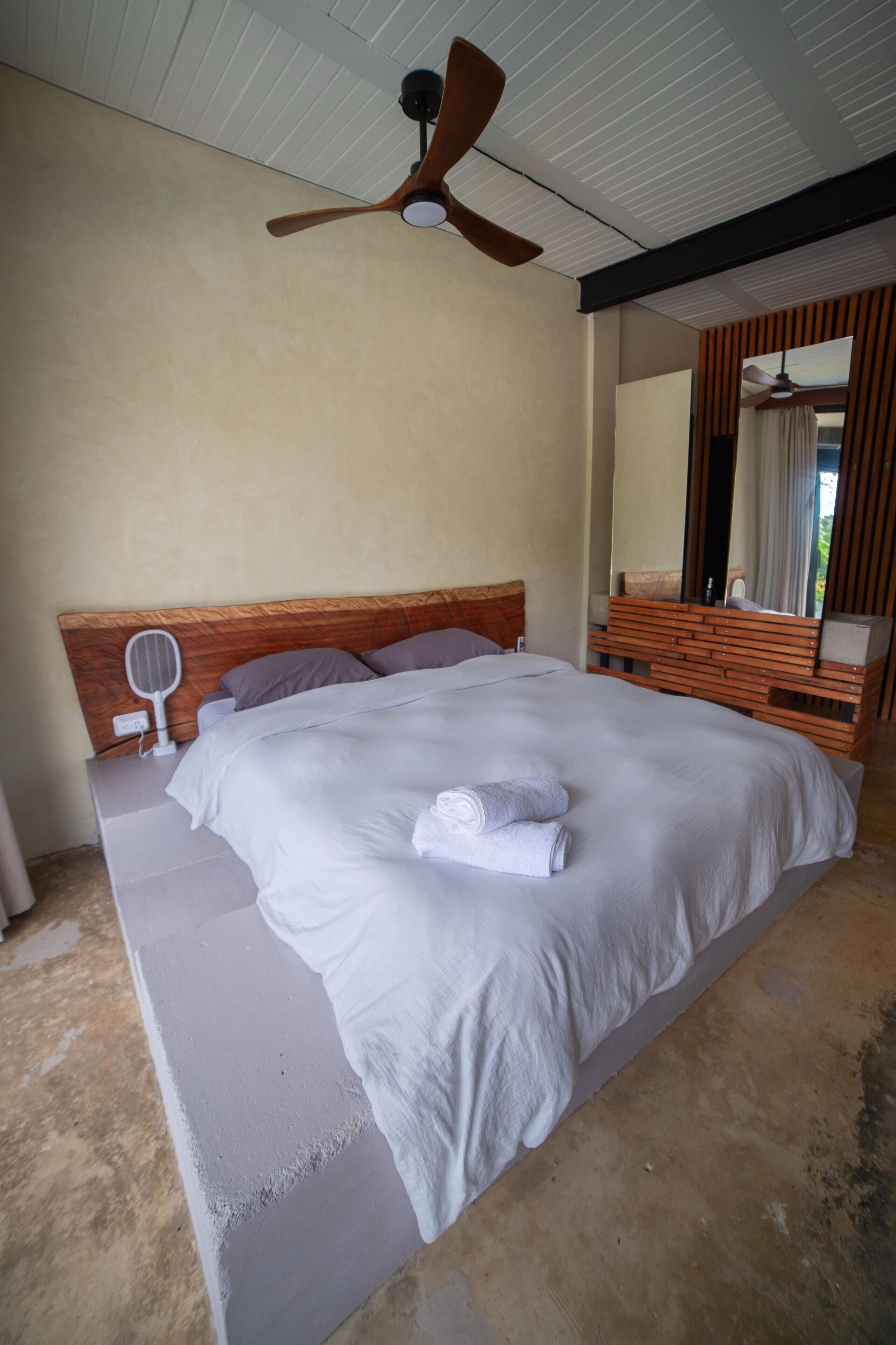 A large bed with a wooden headboard, a ceiling fan, a bedside table with a mirror, and a window with curtains.