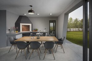 A modern outdoor kitchen with a dining table and chairs, a brick fireplace, and a view of the garden. - Luxury Villa Moon with Pool (Vodnjan)