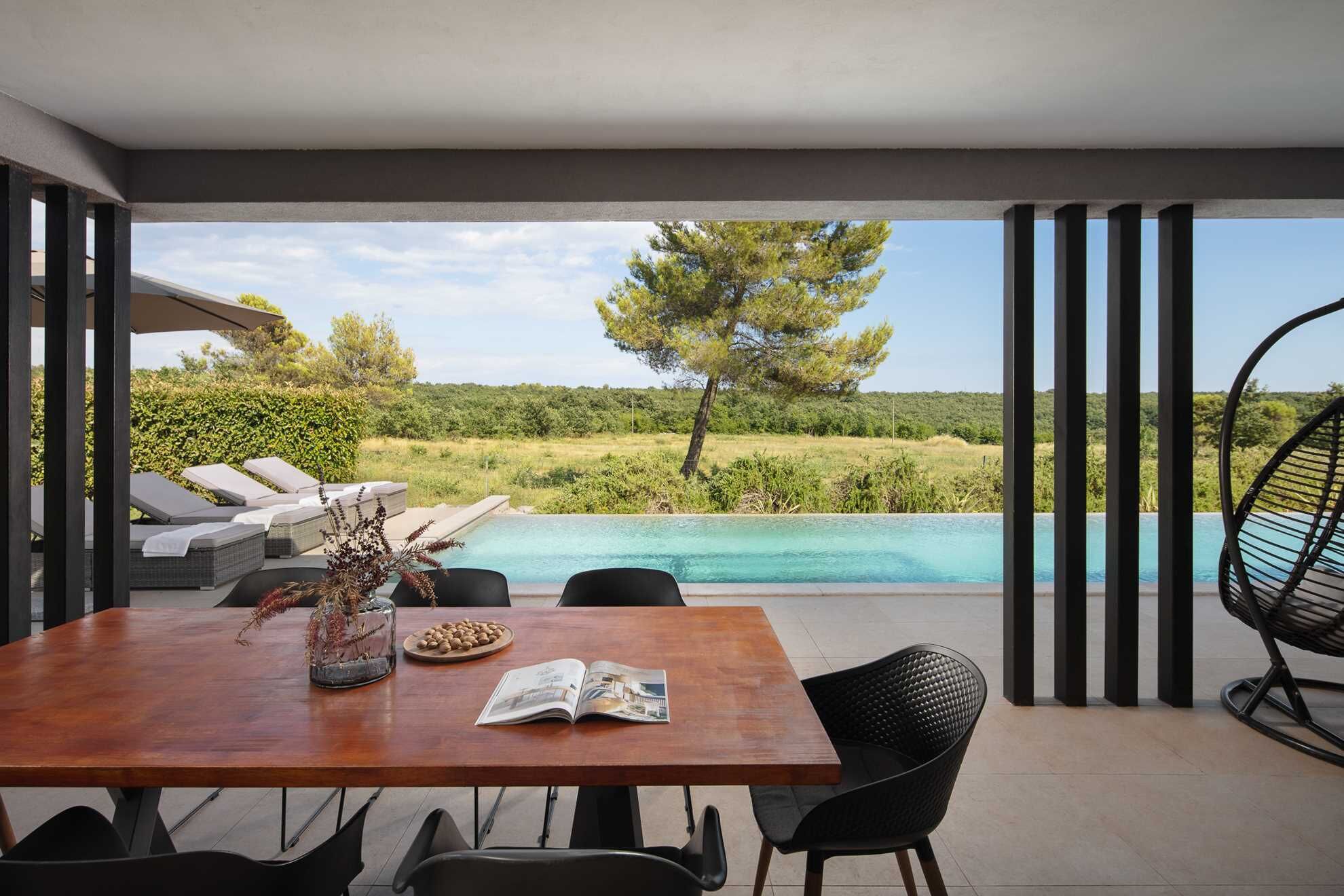 Een moderne eethoek buiten met uitzicht op het zwembad, met een houten tafel, stoelen en een glazen vaas met gedroogde planten.