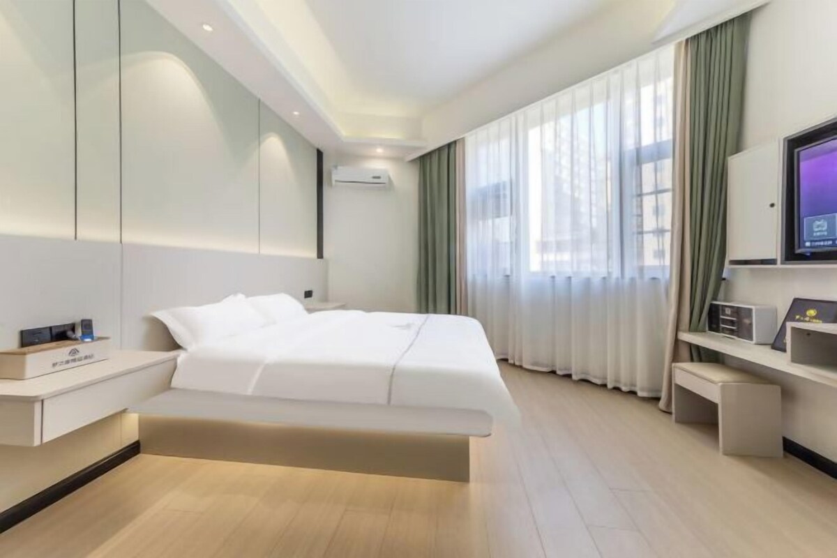A modern hotel room with a large bed, a flat-screen TV, a bedside table, and a window with sheer curtains.