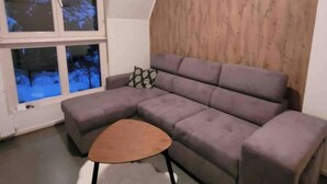 A modern living room with a grey sectional sofa, a wooden coffee table, and a window with a view of trees. - Chalet-style house in the heart of nature (Kruth)