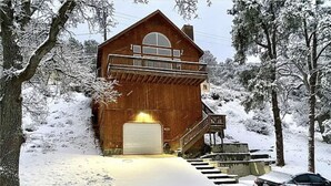 Puinen mökki, jossa on autotalli, ja jonka ympärillä on lumipeitteisiä puita.