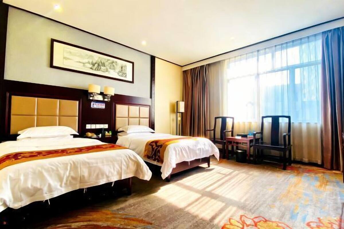 Habitación de hotel con dos camas, un escritorio, una silla, una lámpara y un ventanal con cortinas.