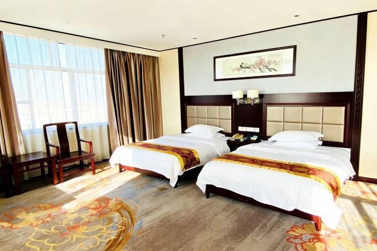 Habitación de hotel con dos camas, un escritorio, una silla y un ventanal grande con cortinas.