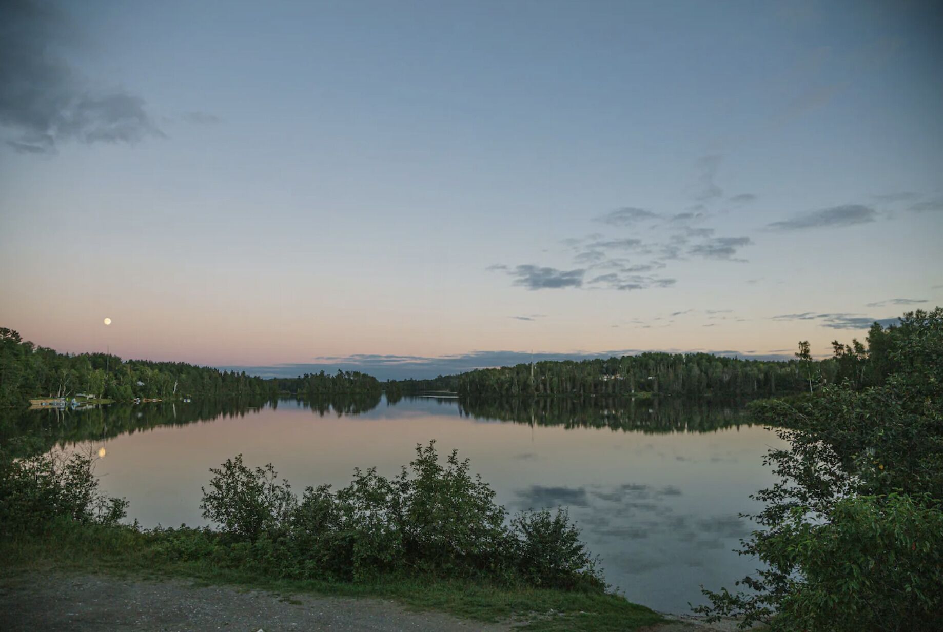 A serene lake at dusk with a clear sky and a visible moon.