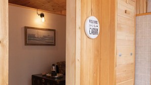 Interior de una cabaña con pared de madera, un cuadro enmarco, una mesita con lámpara y un letrero que dice 'Welcome to the Cabin'.