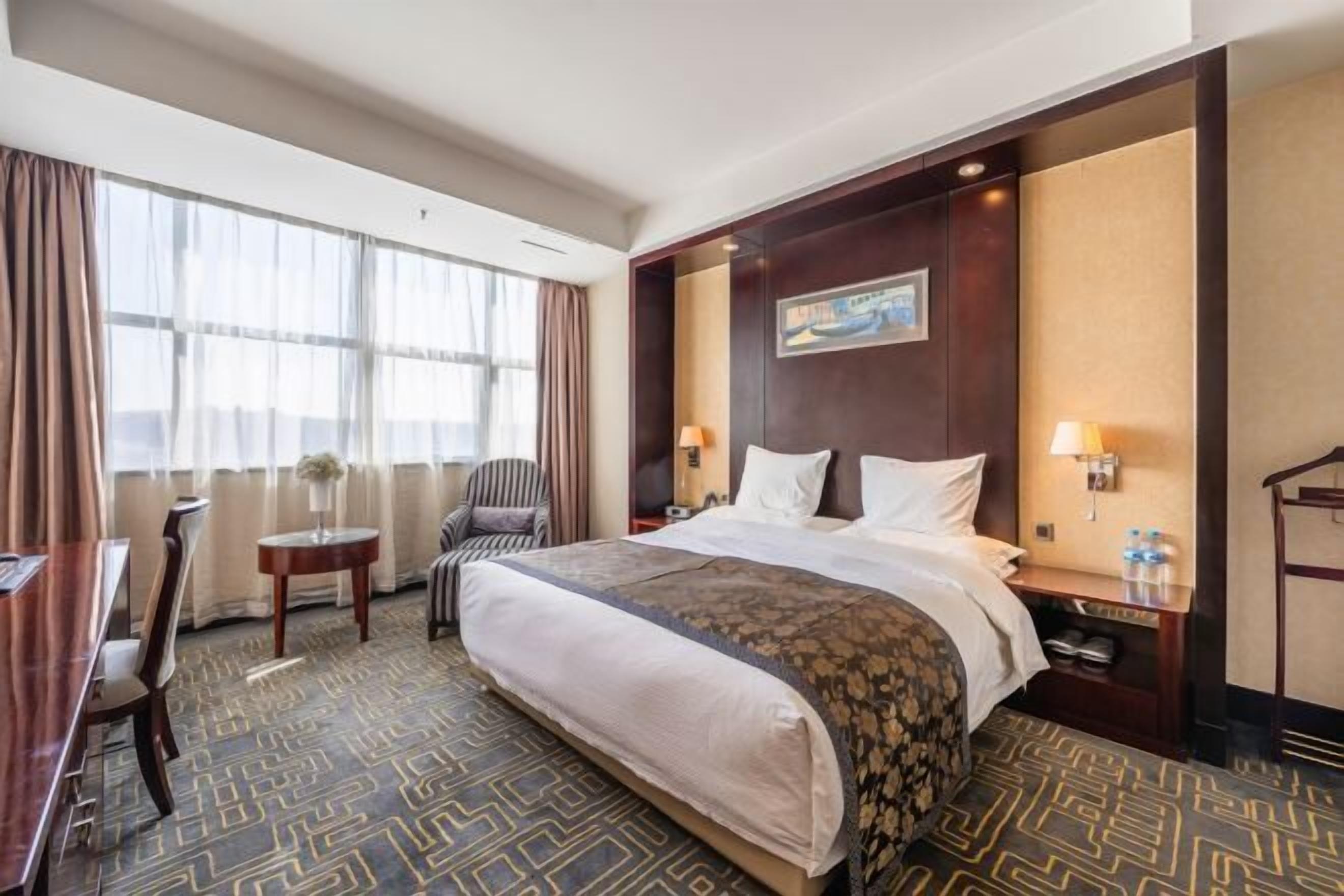 A hotel room with a large bed, a desk, a chair, a small table, and a painting on the wall.