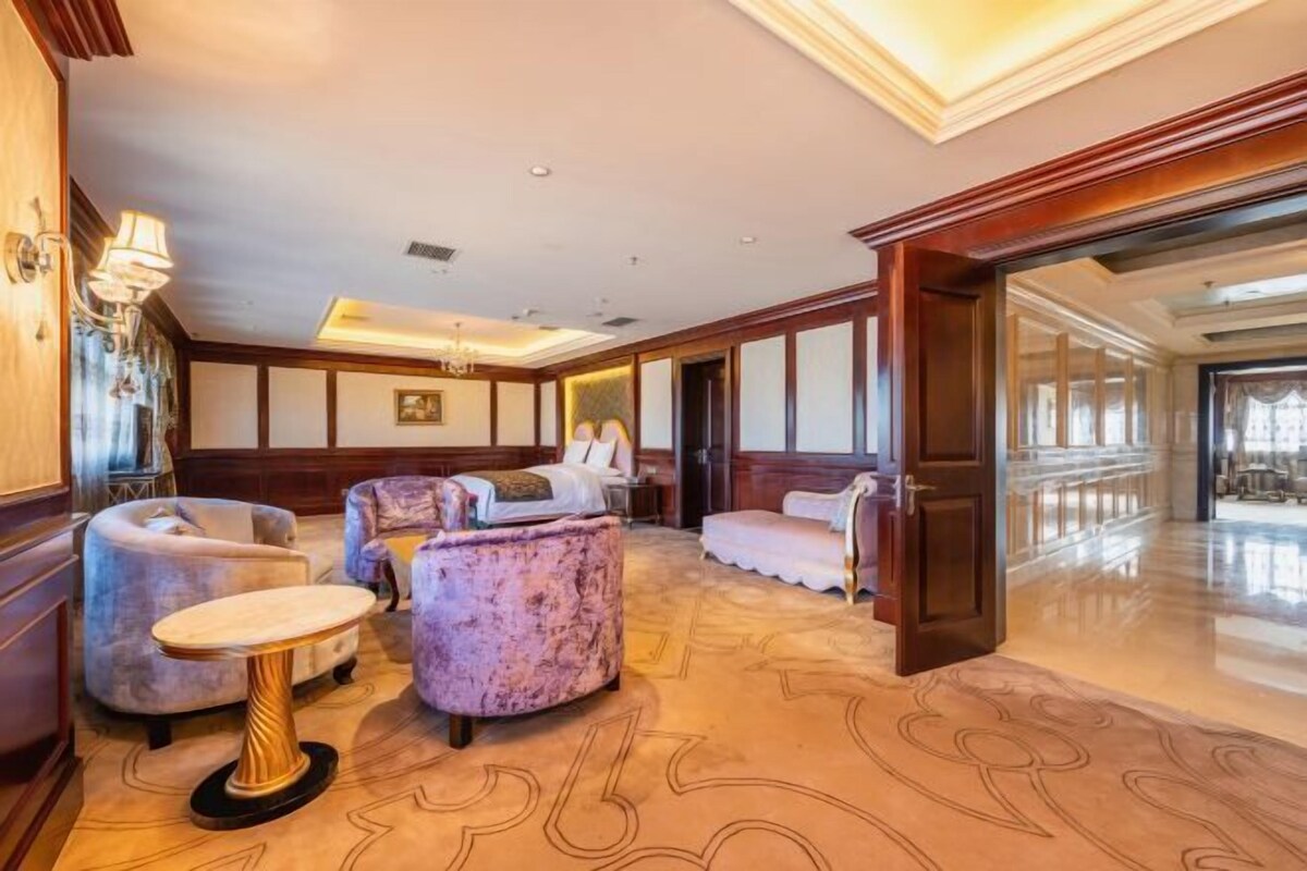 A spacious lobby with a high ceiling, a chandelier, and a patterned carpet. There are plush seating areas and a large window providing natural light.