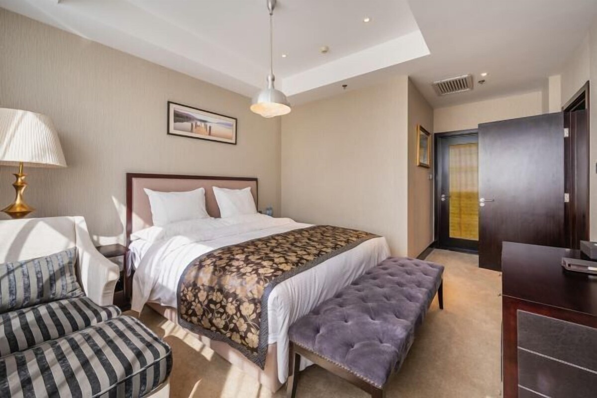 A hotel room with a bed, bedside table, lamp, chair, bench, and wardrobe.