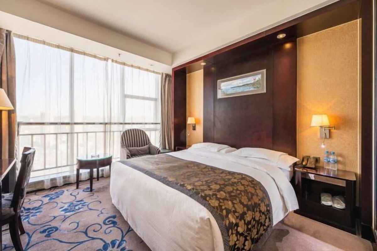 A hotel room with a large bed, a desk, a chair, a window with curtains, and a nightstand with a lamp and a phone.