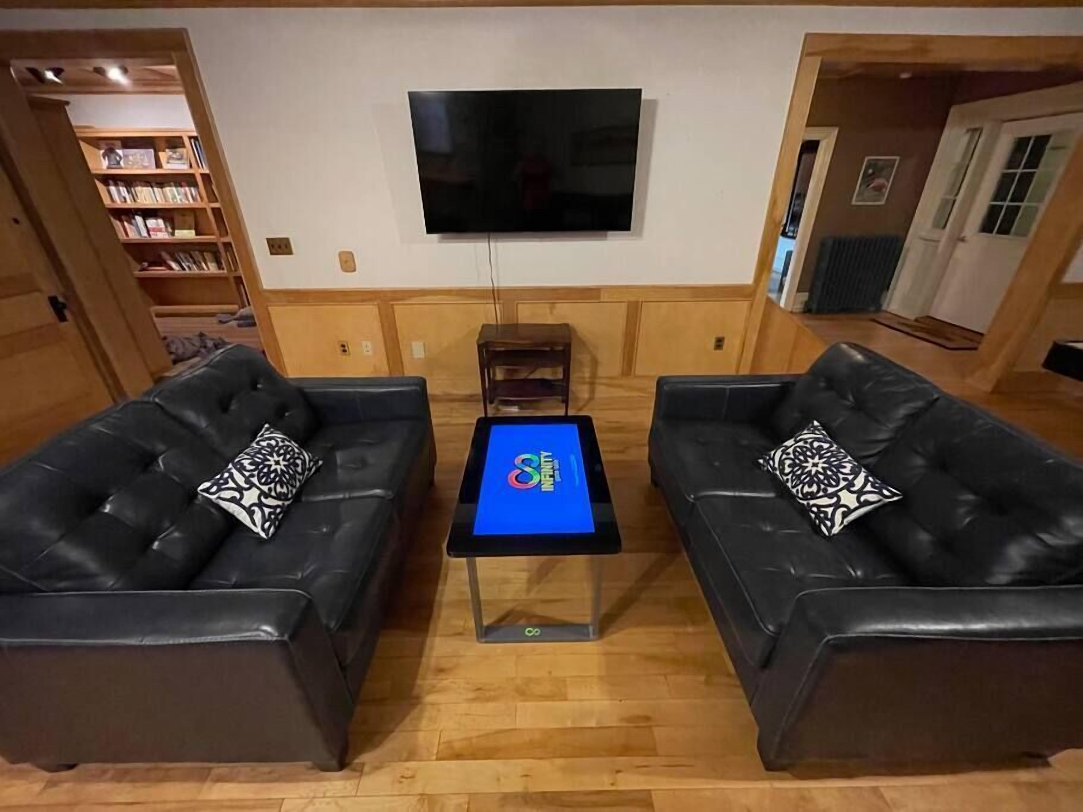 A living room with two black leather sofas, a coffee table with a screen, a flat-screen TV mounted on the wall, and wooden paneling on the walls.