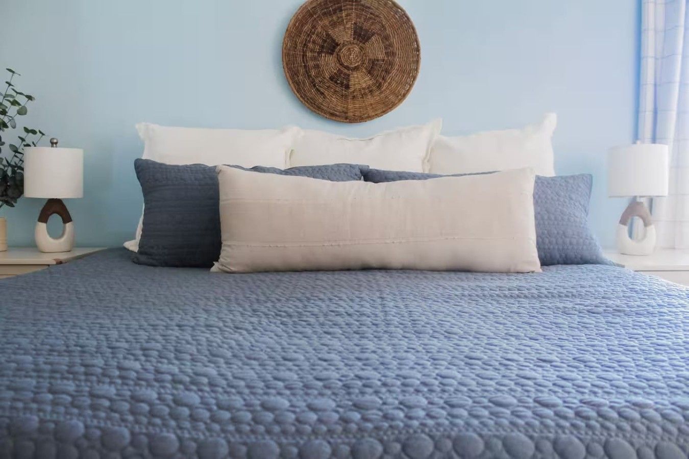 A bed with a textured blue quilt, a round woven wall hanging, and a bedside lamp.