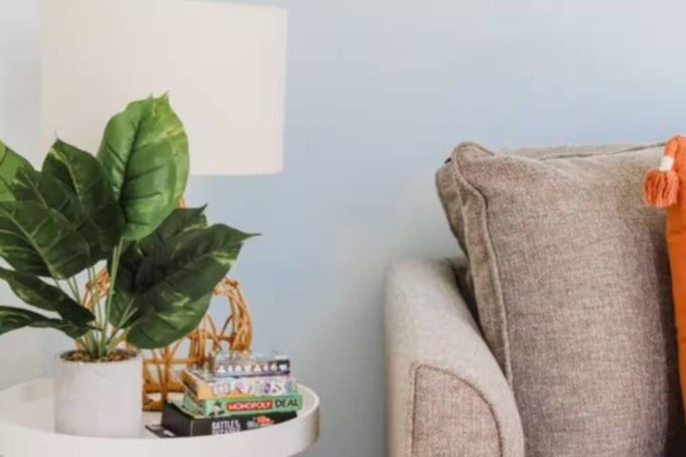 A room with a grey sofa, a white side table with a plant and board games, and a grey cushion with an orange tassel.