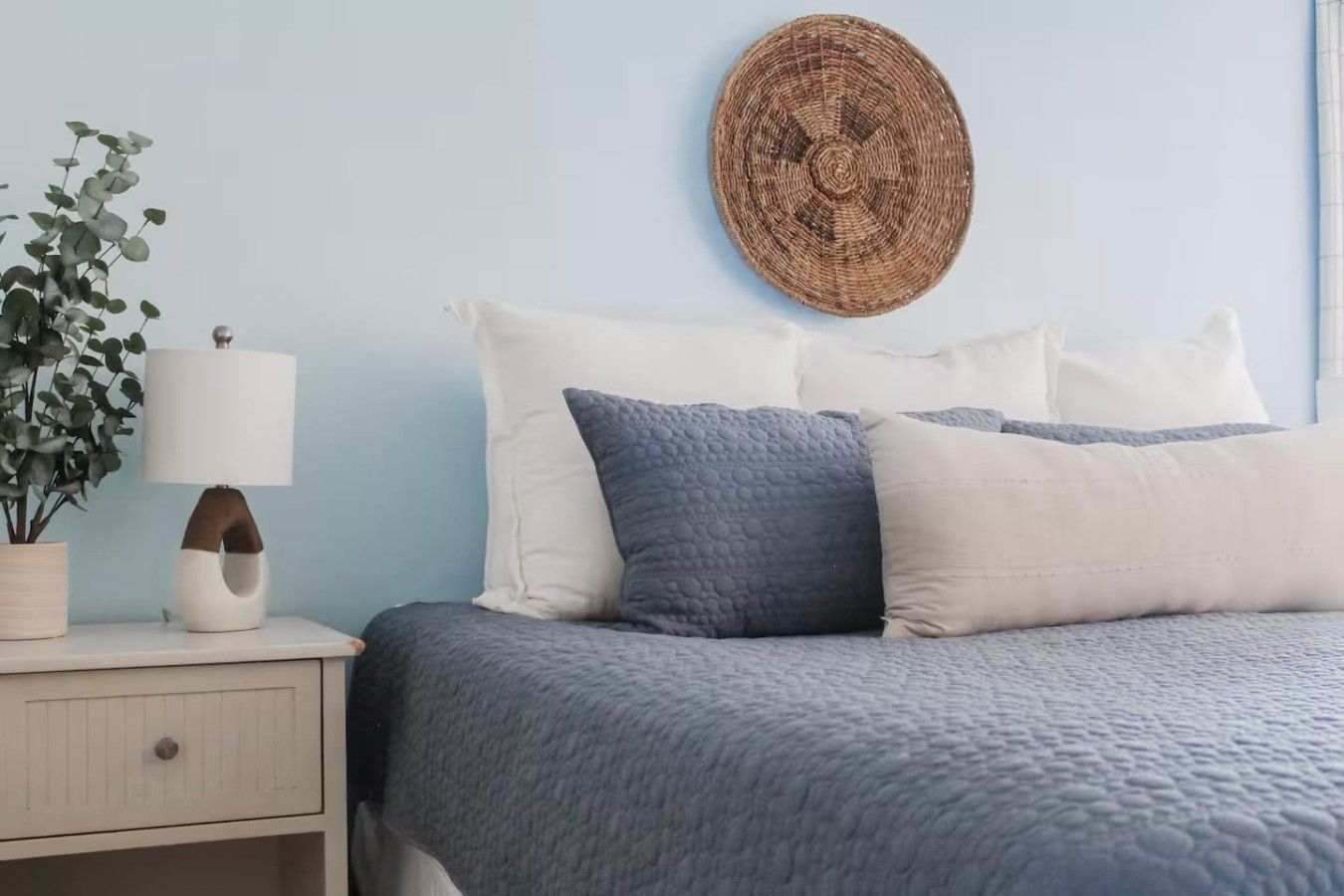 A bed with a blue quilt, white pillows, and a round woven wall hanging.