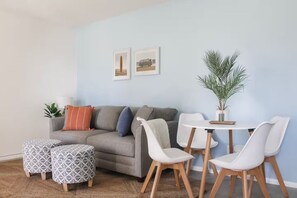 Sala de estar com sofá cinza, pufes estampados, mesa branca com cadeiras e planta em vaso.