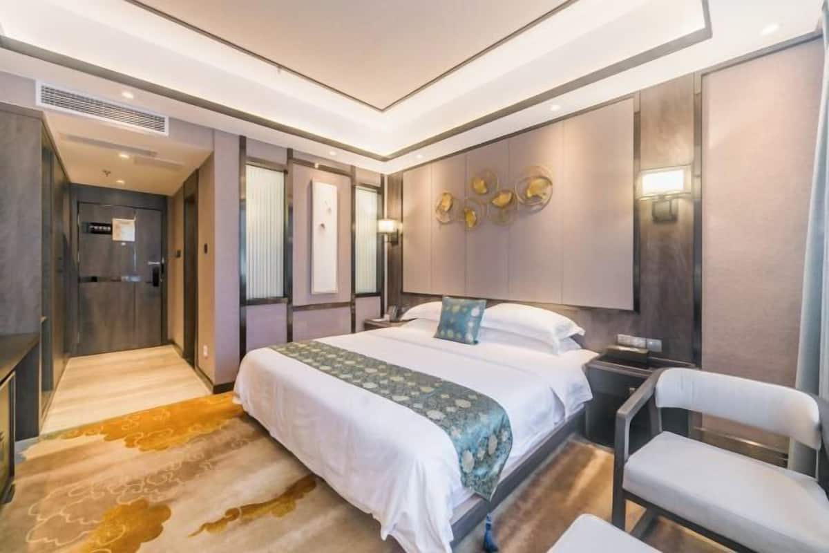 A modern hotel room with a large bed, a desk, and a chair. There is a walk-in closet and a bathroom visible through the door.
