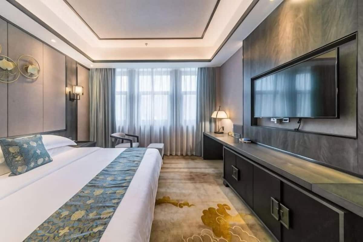 A hotel room with a large bed, a TV mounted on the wall, a desk, and a window with curtains.