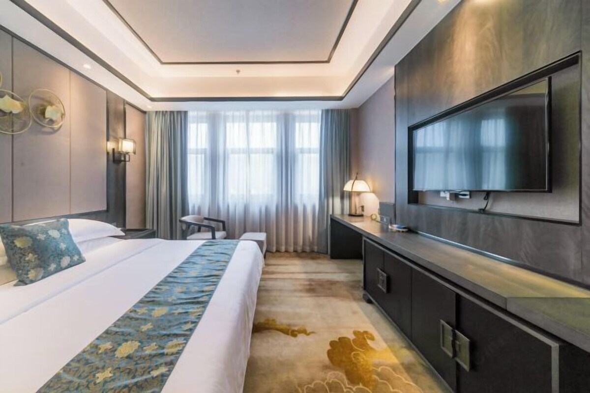 A modern hotel room with a large bed, a flat-screen TV, a desk, and a large window with curtains.