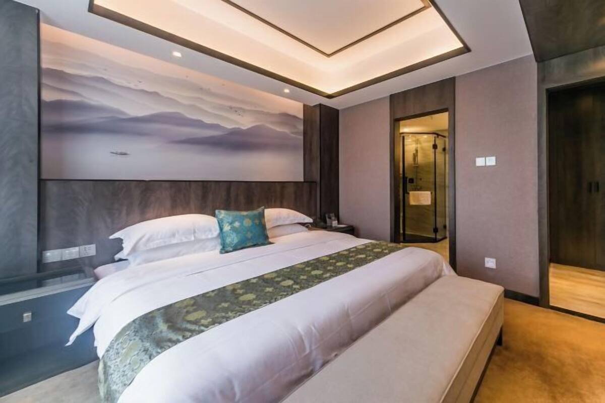 A modern hotel room with a large bed, a headboard with a mural, a bench, and a door leading to a bathroom.